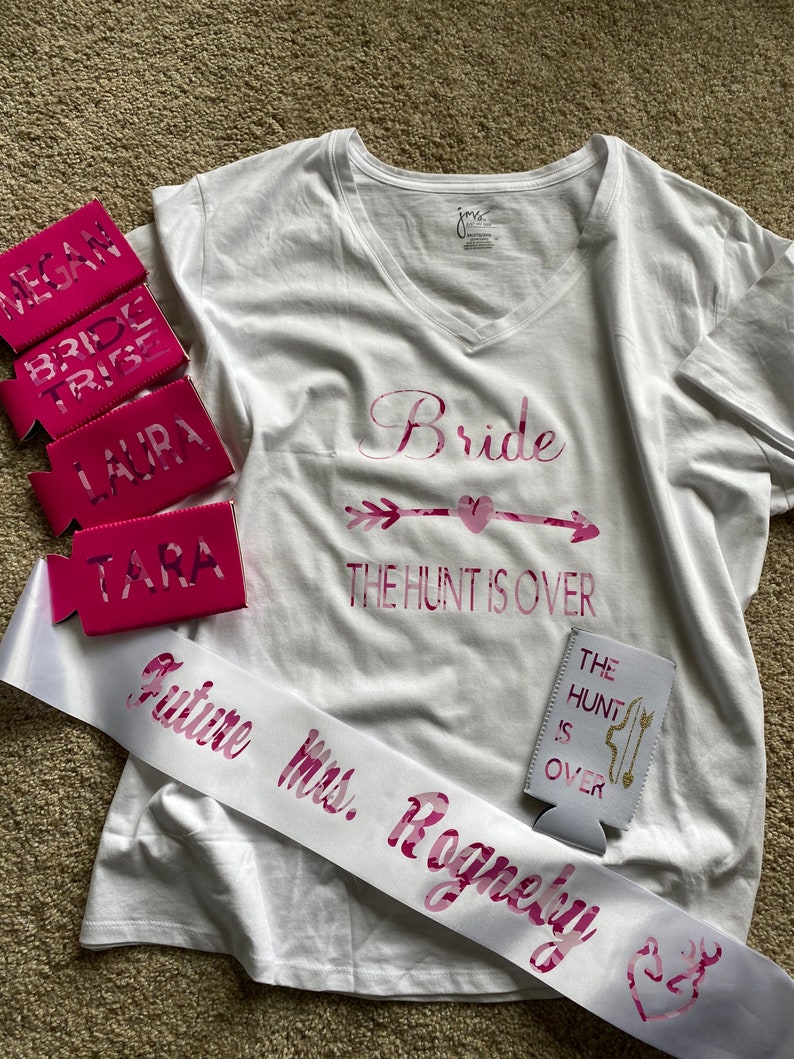 May include: White V-neck t-shirt with "Bride" and "The Hunt Is Over" in pink lettering. Includes pink can coolers with names and "Bride Tribe". A white sash reads "Future Mrs. Rogneby".