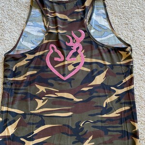 May include: Camouflage tank top with a pink glitter heart-shaped logo. The tank top features a racerback design and is patterned with shades of green, brown, and tan. The logo is a stylized deer head.