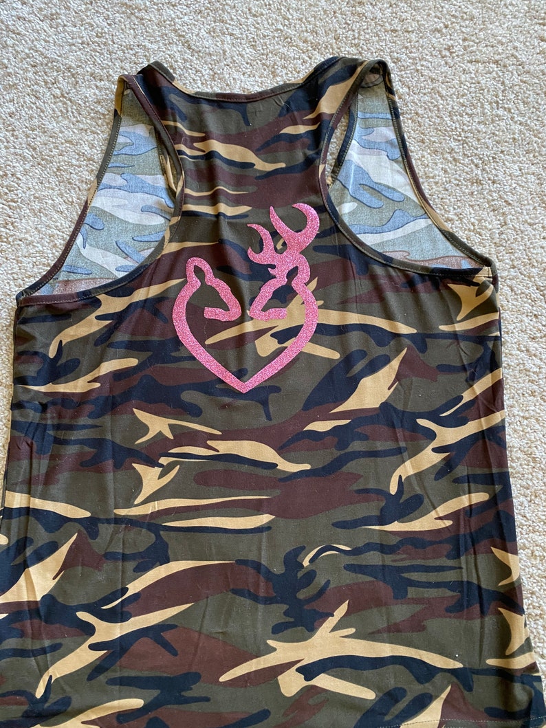 May include: Camouflage tank top with a pink glitter heart-shaped logo. The tank top features a racerback design and is patterned with shades of green, brown, and tan. The logo is a stylized deer head.