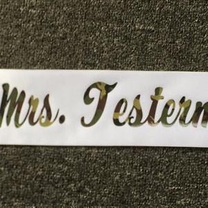 May include: A white satin sash with the text "Future Mrs. Testerman" in a camouflage pattern. The sash is unadorned and appears to be new. The text is in a cursive font.
