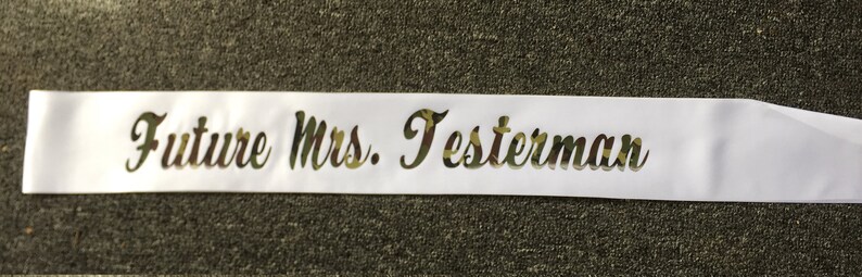 May include: A white satin sash with the text "Future Mrs. Testerman" in a camouflage pattern. The sash is unadorned and appears to be new. The text is in a cursive font.