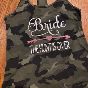 May include: Camouflage print tank top with a racerback design. The text "Bride" is displayed in white script above a pink glitter arrow and heart, with the words "THE HUNT IS OVER" below.