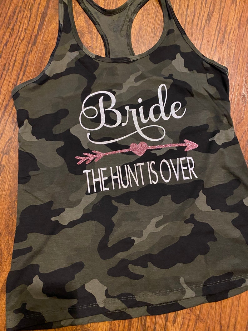 May include: Camouflage print tank top with a racerback design. The text "Bride" is displayed in white script above a pink glitter arrow and heart, with the words "THE HUNT IS OVER" below.