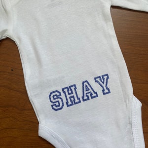 May include: White long-sleeve bodysuit with the name "SHAY" in blue glitter lettering on the back.