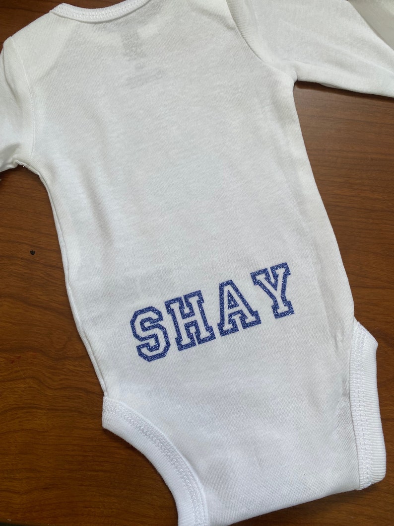 May include: White long-sleeve bodysuit with the name "SHAY" in blue glitter lettering on the back.