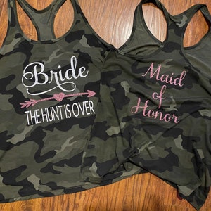 May include: Two camouflage print tank tops. One says "Bride" with an arrow and the text "THE HUNT IS OVER" in white. The other says "Maid of Honor" in pink glitter lettering. Both tops have a racerback design.