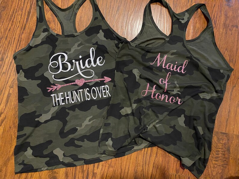 May include: Two camouflage print tank tops. One says "Bride" with an arrow and the text "THE HUNT IS OVER" in white. The other says "Maid of Honor" in pink glitter lettering. Both tops have a racerback design.