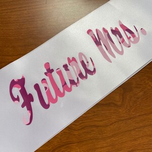 May include: White satin sash with the text "Future Mrs." in a pink camouflage pattern. The sash is angled across a wooden surface, showcasing the elegant script and color scheme.