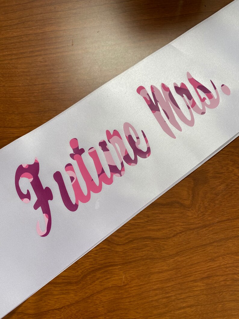 May include: White satin sash with the text "Future Mrs." in a pink camouflage pattern. The sash is angled across a wooden surface, showcasing the elegant script and color scheme.