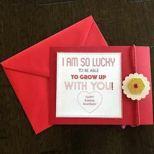 May include: A red greeting card and envelope set. The card reads "I AM SO LUCKY TO BE ABLE TO GROW UP WITH YOU! HAPPY RAKSHA BHANDAN!" A decorative flower with a gold center is attached to the card.