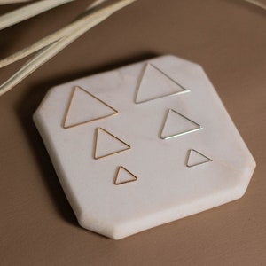 May include: Five different sized gold and silver triangle hoop earrings displayed on a white marble surface.