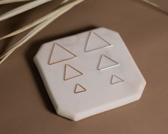 May include: Five different sized gold and silver triangle hoop earrings displayed on a white marble surface.
