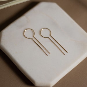 May include: A pair of gold-toned earrings with a simple circle design. The earrings are on a white marble surface.