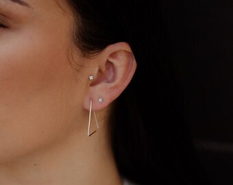 May include: A person wearing a silver triangle hoop earring and two small silver stud earrings in their ear.