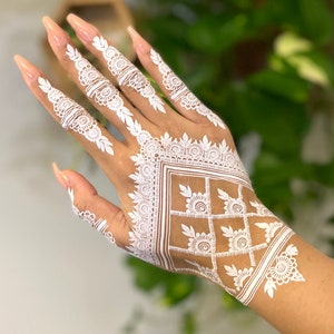 May include: A hand with white henna designs on the back of the hand and fingers. The henna is intricate and detailed, with a geometric pattern on the back of the hand and floral designs on the fingers.