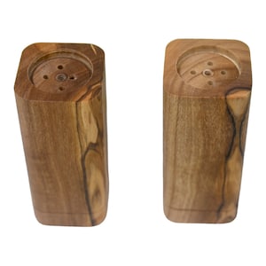 May include: A set of two wooden salt and pepper shakers with a natural wood finish. The shakers are square with rounded edges and have a small hole in the top for dispensing salt and pepper.