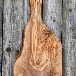 May include: An olive wood chopping board with a natural edge and a handle. The board is a warm brown colour with distinctive grain patterns. It has a hole in the handle for hanging. The background is a weathered wooden wall.