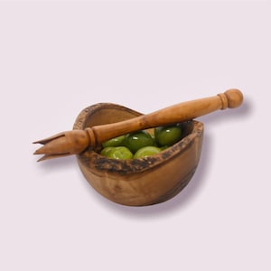 May include: A small wooden bowl with a carved handle, filled with green olives. The bowl is made of olive wood and has a natural, rustic look.