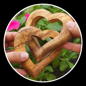 May include: Two intricately carved wooden hearts, one nested inside the other, are held in the hands of a person. The hearts are made of olive wood and have a natural brown color with visible grain patterns.