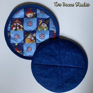May include: Two round, quilted pot holders. One pot holder features a blue and white checkered pattern with snowmen and snowflakes. The other pot holder is solid blue. The pot holders have a blue binding. The text "Two Bears Studio" is in the upper right corner.