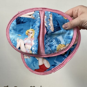 May include: Two pink pot holders with a blue and white fabric featuring a vintage pin-up girl design. The fabric has a blue and white scalloped trim.