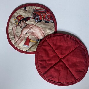 May include: Two round pot holders. One features a firefighter graphic with a red fire truck and an axe. The other is solid red with a quilted pattern. Both have a red border.