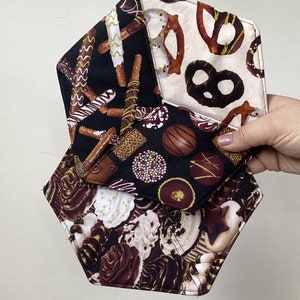 May include: Hexagon-shaped fabric items with various chocolate and pretzel patterns. The fabrics feature designs of chocolate sweets, pretzels, and other treats in shades of brown, white, and black. The items are held by a hand.