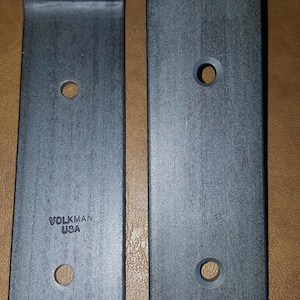 May include: Two metal brackets with holes and a rounded edge. The bracket on the left has the text "VOLKMAN USA" printed on it.