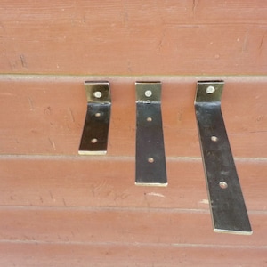May include: Three metal shelf brackets in different sizes. The brackets are made of black metal and have a slightly curved shape. They are mounted on a wooden wall.