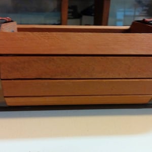 Op de afbeelding: Een houten plantenbak met metalen banden. De plantenbak is gemaakt van lichtbruin hout en heeft een rechthoekige vorm. De metalen banden zijn zilverkleurig en zijn met schroeven aan de plantenbak bevestigd.