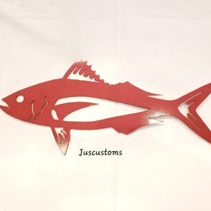 May include: A red metal fish silhouette with a detailed fin and tail design. The fish has a cut-out design and is mounted on a white background. The text "Juscustoms" is visible on the lower right side. This decorative piece is suitable for nautical or coastal decor.