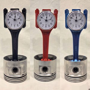 May include: Three clocks with white faces and black hands, set in a piston and connecting rod base. The connecting rods are painted in black, red, and blue. The clock faces have black numbers and a quartz label.