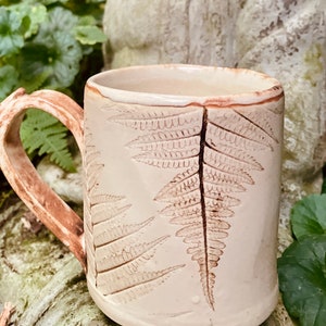 May include: A ceramic mug with a light brown glaze and a fern design. The mug has a large handle and is perfect for enjoying a hot beverage.