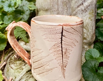 May include: A ceramic mug with a light brown glaze and a fern design. The mug has a large handle and is perfect for enjoying a hot beverage.