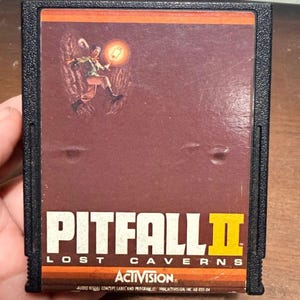 May include: A black rectangular video game cartridge for the Atari 2600 featuring a brown label with the title Pitfall II Lost Caverns in white and yellow text. The label displays an illustration of a character holding a lantern in a cavern.