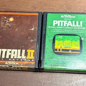 May include: Two rectangular black plastic video game cartridges for the Atari system. The left cartridge is labeled Pitfall II Lost Caverns by Activision. The right cartridge is green and labeled Pitfall! Pitfall Harry's Jungle Adventure by Activision.