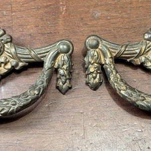 May include: Two antique brass drawer pulls with ornate floral and leaf designs. Each pull features a curved handle and decorative accents, showcasing a vintage aesthetic. The pulls are approximately 6 inches wide.