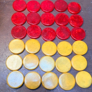 May include: A collection of thirty circular game pieces arranged in six rows of five. The top three rows consist of fifteen red marbled discs, while the bottom three rows feature fifteen yellow marbled discs, all with a smooth, flat surface.