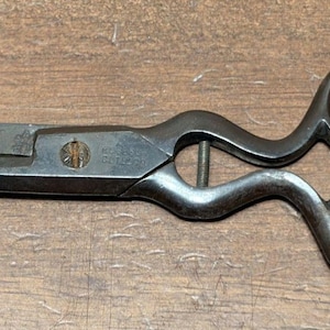 May include: Antique metal scissors with a curved handle design and rounded finger holes. The scissors have a pointed blade and a dark, aged finish, suggesting vintage craftsmanship. The scissors are resting on a wooden surface.