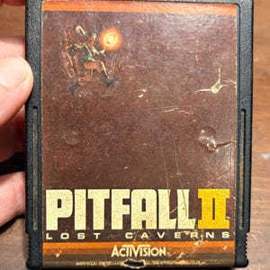 May include: A black rectangular video game cartridge for the Atari 2600 featuring a brown label with the title Pitfall II Lost Caverns in yellow and white text. The label displays an illustration of a character holding a glowing lantern.