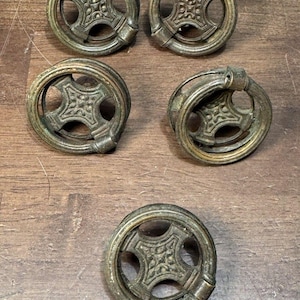 May include: A collection of five antique brass drawer pulls. Each pull features a circular design with a decorative center and a hinged handle. The brass has a weathered, aged appearance, suggesting vintage or reclaimed hardware.