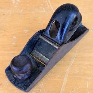 May include: A vintage Stanley block plane featuring a dark metal body with a worn finish, a rounded wooden front knob, and a curved black lever cap. The tool includes a metallic blade mechanism and shows signs of age with surface oxidation.