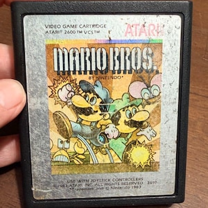 May include: A black Atari 2600 video game cartridge for Mario Bros. by Nintendo. The silver label features a colorful illustration of Mario and Luigi, with text reading VIDEO GAME CARTRIDGE, ATARI 2600 TM VCS TM, MARIO BROS. BY NINTENDO, and 1983.