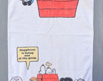 May include: White rectangular towel featuring Snoopy and the Peanuts gang. Snoopy is hanging upside down from his red doghouse. The towel has the phrase "Happiness is being one of the gang." at the bottom, with the Peanuts characters.