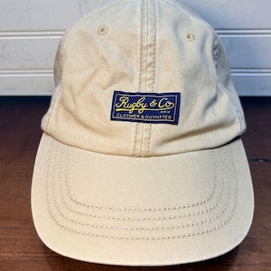 May include: A beige baseball cap with a structured crown and a curved brim. Centered on the front is a rectangular navy blue patch featuring the yellow embroidered text Rugby & Co CLOTHIER & OUTFITTER. The cap includes matching beige stitching.