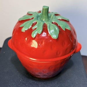 May include: A glossy red ceramic lidded container shaped like a strawberry. The lid features a sculpted green stem and leaves at the top. The body of the container has a textured surface with small indentations and two small side handles.