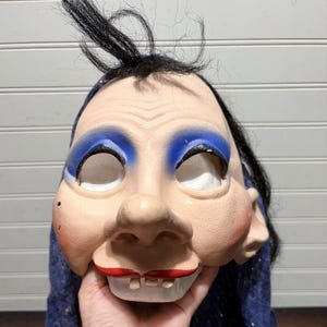 May include: A full-head mask featuring a pale, exaggerated facial expression with bright blue eyeshadow, rosy cheeks, and red lips. The mask has prominent front teeth, a large nose, and sparse black hair attached to the top and sides.