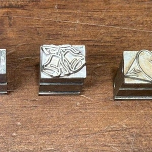May include: Five metal stamps with various designs, including the letters "M", a heart pierced by an arrow, and a pentagram. The stamps are rectangular and appear to be made of a silver-colored metal. They are arranged in a row on a wooden surface.