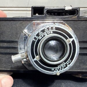 May include: Vintage Argus camera with a black body and a silver lens. The lens has a black and white dial with numbers and the word "Argus". The camera has a silver winding knob and a silver button on the side.