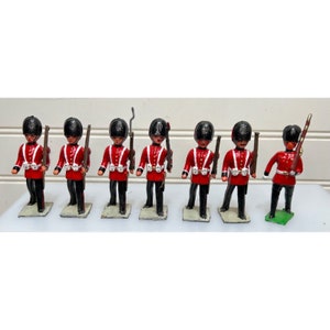 May include: A collection of seven toy soldiers in the style of the Queen's Guard. Each figure is dressed in a red tunic, black trousers, and a tall black hat. They are holding rifles and standing on small bases.